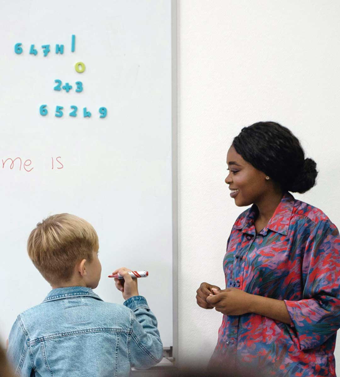 face-face-1080×1200 Teacher assisting young student at whiteboard with math problems in a classroom setting